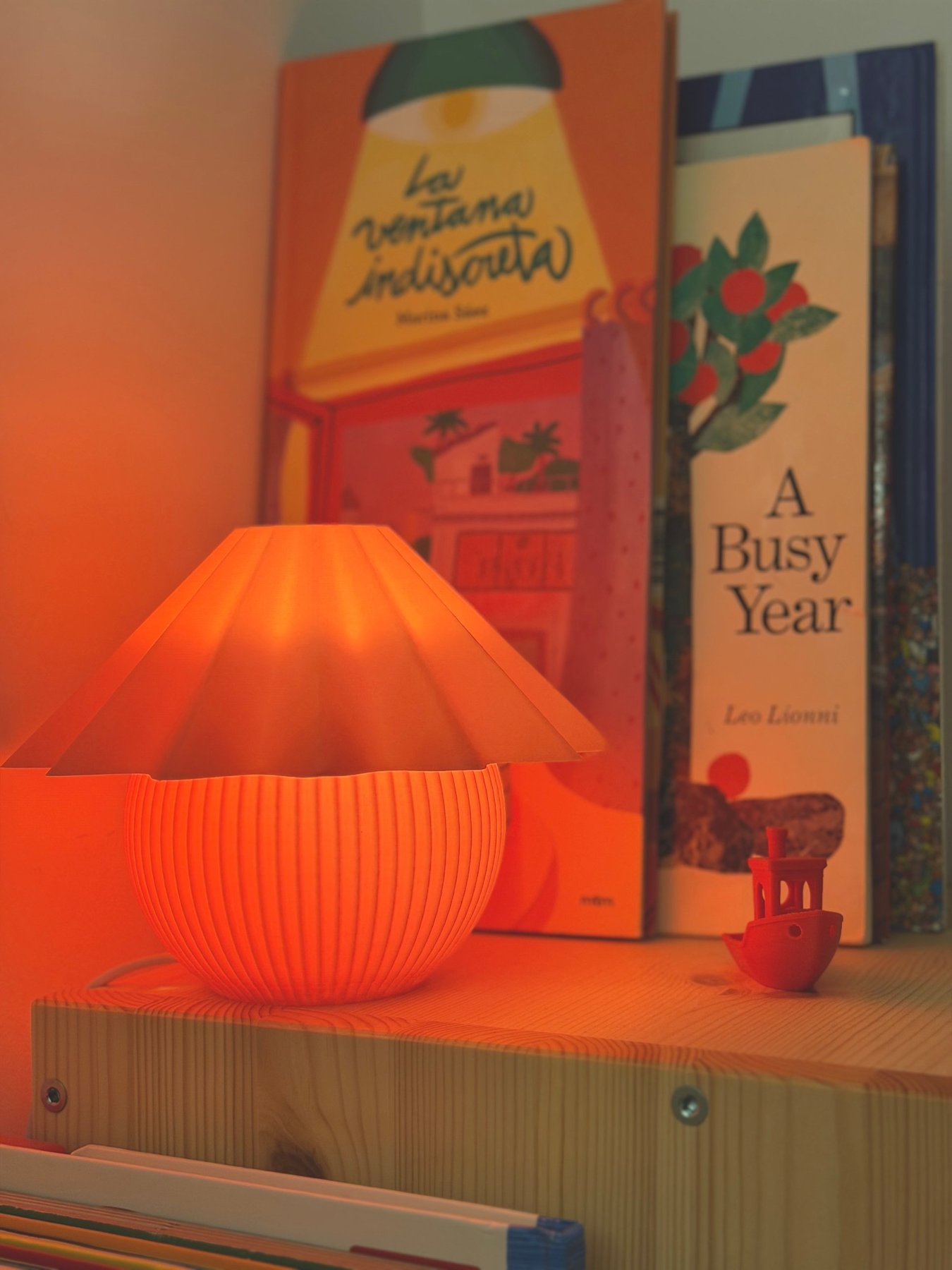 Penélope naranja encendida en una estantería, junto a libros ilustrados y un barquito impreso en 3D